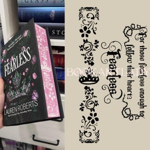May include: A hardcover book titled "FEARLESS" by Lauren Roberts, with a black cover featuring a crown and pink accents. The book's edges are pink with a white floral pattern. Decorative black floral elements and text are on a beige background.