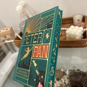 May include: A green hardcover book with gold lettering and illustrations. The title is "Peter Pan" by J.M. Barrie. The book has a decorative border with fairies and other whimsical elements.