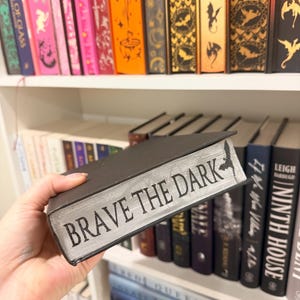 Pu&ograve; includere: Un libro nero con il titolo "BRAVE THE DARK" in lettere argentate. Il libro &egrave; tenuto davanti a una libreria piena di vari libri, molti con dorsi decorativi. La mensola &egrave; bianca e i libri sono di diversi colori e dimensioni.