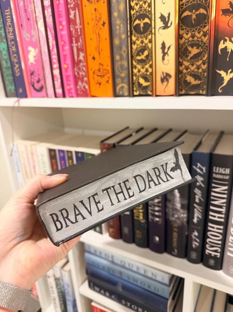 Pu&ograve; includere: Un libro nero con il titolo "BRAVE THE DARK" in lettere argentate. Il libro &egrave; tenuto davanti a una libreria piena di vari libri, molti con dorsi decorativi. La mensola &egrave; bianca e i libri sono di diversi colori e dimensioni.