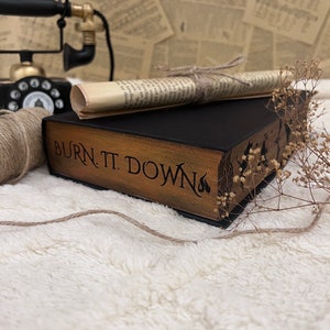Pode incluir: Um livro preto com capa de madeira e o t&iacute;tulo "BURN. T. DOWN" em letras douradas. O livro est&aacute; sobre uma superf&iacute;cie branca e fofa com um peda&ccedil;o de papel enrolado e um telefone rotativo vintage no fundo.