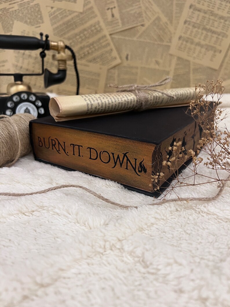 Pode incluir: Um livro preto com capa de madeira e o t&iacute;tulo "BURN. T. DOWN" em letras douradas. O livro est&aacute; sobre uma superf&iacute;cie branca e fofa com um peda&ccedil;o de papel enrolado e um telefone rotativo vintage no fundo.