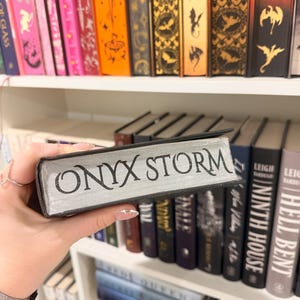 Pu&ograve; includere: Un libro intitolato "ONYX STORM" con lettere argentate &egrave; tenuto davanti a una libreria piena di libri diversi. I dorsi dei libri sullo scaffale sono di vari colori, tra cui rosa, arancione e nero.