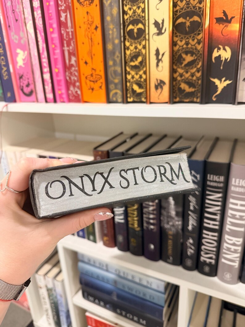 Pu&ograve; includere: Un libro intitolato "ONYX STORM" con lettere argentate &egrave; tenuto davanti a una libreria piena di libri diversi. I dorsi dei libri sullo scaffale sono di vari colori, tra cui rosa, arancione e nero.
