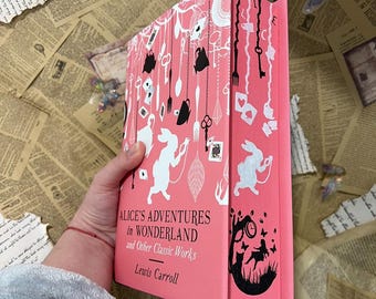 Puede incluir: Un libro de tapa dura rosa con una ilustración en blanco y negro de Alicia en el País de las Maravillas en la portada. El título es "Alice's Adventures in Wonderland and Other Classic Works" de Lewis Carroll.