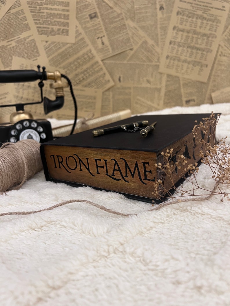 Pode incluir: Uma caixa de madeira com uma tampa de couro preto e as palavras "IRON FLAME" inscritas na frente. A caixa est&aacute; sobre uma superf&iacute;cie branca e fofa com uma pequena faca prateada e um peda&ccedil;o de barbante.