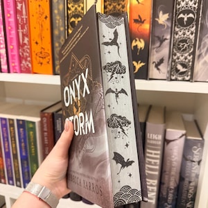 Pu&ograve; includere: Un libro con il titolo "Onyx Storm" di Rebecca Yarros, tenuto davanti a una libreria. Il libro ha una copertina scura con dettagli dorati e il titolo in bianco. Il dorso presenta illustrazioni di draghi e nuvole.