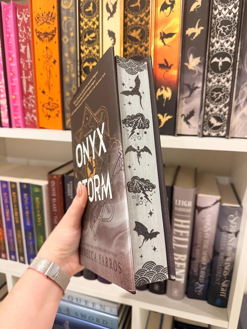 Pu&ograve; includere: Un libro con il titolo "Onyx Storm" di Rebecca Yarros, tenuto davanti a una libreria. Il libro ha una copertina scura con dettagli dorati e il titolo in bianco. Il dorso presenta illustrazioni di draghi e nuvole.