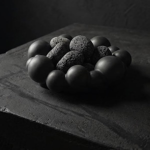 Könnte beinhalten: Eine schwarze dekorative Schale mit einem abgerundeten, blasenartigen Rand, die mehrere strukturierte, dunkelgraue Steine enthält. Die Schale und die Steine ​​sind vor einem dunklen Hintergrund platziert und erzeugen eine monochrome, minimalistische Ästhetik.