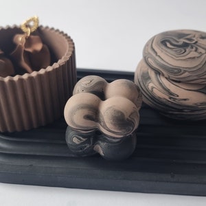 May include: A decorative arrangement featuring a brown, ribbed container holding brown fabric, a black tray, and two marbled stone objects. The stone objects have a beige and black swirl pattern. The tray has a curved design.