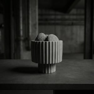 Puede incluir: Un cuenco de hormigón gris acanalado con tres piedras grises texturizadas. El cuenco tiene forma cilíndrica con ranuras verticales y se asienta sobre un pedestal escalonado corto. El fondo es un entorno industrial oscuro.