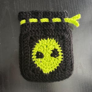 Puede incluir: Una bolsa negra de ganchillo con un diseño de cara de alienígena verde lima en la parte delantera. La bolsa tiene un cierre de cordón y un acento verde lima alrededor de la parte superior. La cara alienígena tiene dos ojos y una boca.