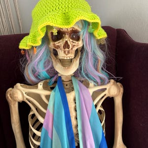 May include: A skeleton wearing a bright green crochet hat with colorful beads, a blue, pink, and green striped scarf, and a long, multi-colored wig.
