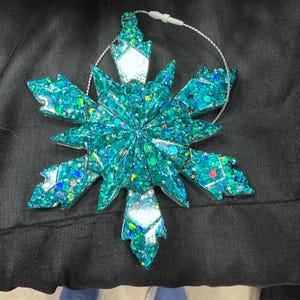 May include: A teal blue snowflake Christmas ornament with glitter. The snowflake has a silver centre and is made of plastic.