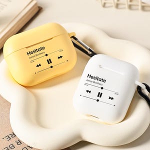 May include: Two wireless earbud cases, one yellow and one white, each with a black carabiner clip. The cases feature the text "Hesitate Jonas Brothers" and a music player graphic. They rest on a cream-coloured, cloud-shaped tray.
