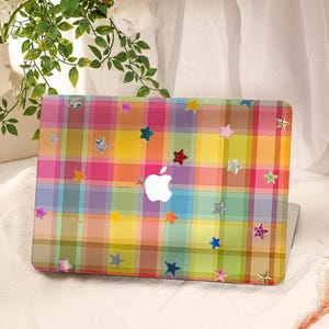 May include: A laptop cover featuring a vibrant, multi-colored plaid pattern in shades of pink, yellow, blue, and green. The surface is decorated with numerous small, scattered star-shaped stickers in various colors, textures, and finishes.