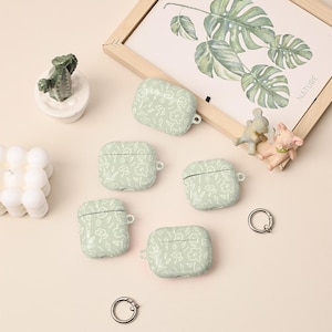 May include: Five pale green AirPod cases, patterned with white flowers, are displayed on a light green surface. A small cactus in a white pot, a framed botanical print, and two small animal figurines are also present. Two silver key rings are also visible.