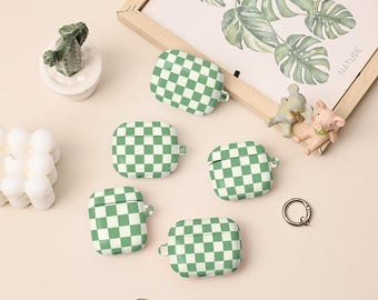 May include: Five white and green checkered pattern earbud cases. The cases are square-shaped with a small loop for a keychain. A small cactus figurine and a framed botanical print are also in the image.