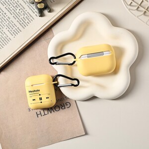 May include: Two yellow AirPod cases with black carabiner clips. One case is open, revealing the white earbuds. The other case has the text "Hesitate Jonas Brothers" and music player controls. The cases are on a white, cloud-shaped tray.
