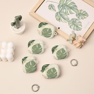 Puede incluir: Cinco fundas blancas para AirPod con un diseño de hoja de monstera verde. Las fundas están dispuestas sobre una superficie beige claro, junto con una impresión botánica enmarcada, pequeñas figuras y llaveros. Las fundas tienen un acabado liso.