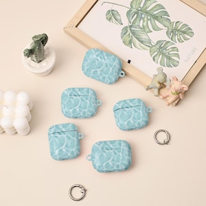 May include: Five light blue AirPod cases with a water ripple design. The cases are arranged on a light beige surface, along with small decorative items, including a framed botanical print and miniature animal figurines.