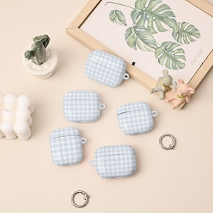 Pode incluir: Cinco capas para AirPods com padrão xadrez vichy azul claro e branco. As capas estão dispostas sobre uma superfície bege clara, juntamente com itens decorativos, incluindo uma impressão botânica emoldurada e figuras de animais em miniatura.