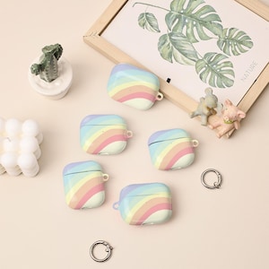 Könnte beinhalten: Eine Sammlung von fünf AirPod-Hüllen mit pastellfarbenen Regenbogenstreifen. Die Hüllen sind weiß mit einem geschwungenen Regenbogenmuster in Rosa, Gelb, Blau und Grün. Eine kleine Kaktusfigur und ein botanischer Druck sind ebenfalls zu sehen.