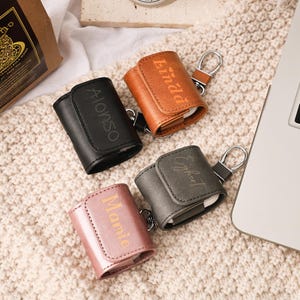 May include: Four personalized leather AirPod cases in black, brown, gray, and pink. Each case has a name engraved on the front: Alonso, Linda, Egbert, and Mamie. The cases have a snap closure and a keychain ring.