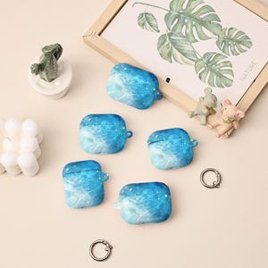 Puede incluir: Se muestran cinco fundas para AirPods con estampado azul y blanco, cada una con un pequeño lazo. También son visibles una pequeña figura de cactus, una impresión botánica enmarcada y dos llaveros. Las fundas tienen un efecto acuarela.