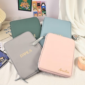Könnte beinhalten: Vier personalisierte Reißverschlusstaschen in Grün, Blau, Grau und Rosa. Jede Tasche hat einen goldfarbenen Namen auf der Vorderseite. Die Taschen haben einen Griff an der Seite und einen Reißverschluss.