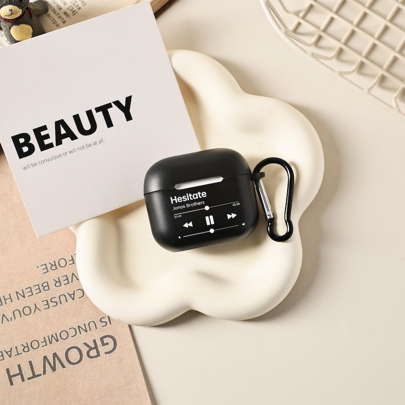 May include: Black AirPod case with a black carabiner clip. The case has the word "Hesitate" and "Jonas Brothers" printed on it, along with a music player interface. A white card with the word "BEAUTY" is in the background.