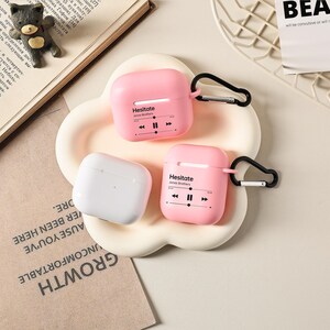 May include: Three AirPod cases in the image. Two are pink with the text "Hesitate" and a music player graphic. The third case is white. All cases have a black carabiner clip attached. The cases are on a white, flower-shaped tray.