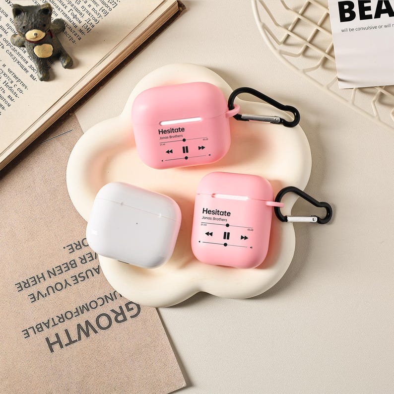 May include: Three AirPod cases in the image. Two are pink with the text "Hesitate" and a music player graphic. The third case is white. All cases have a black carabiner clip attached. The cases are on a white, flower-shaped tray.