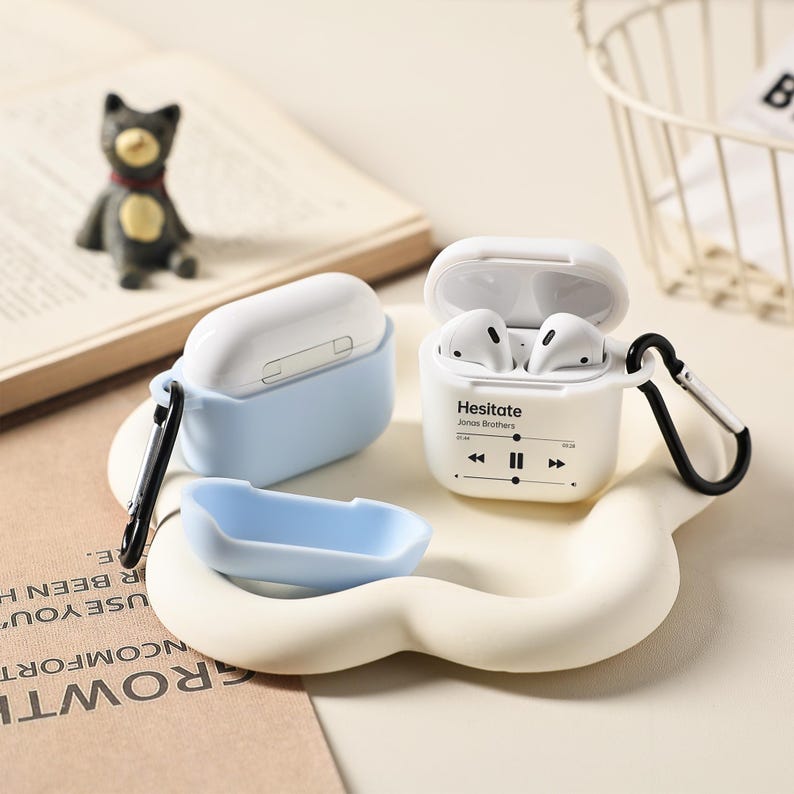 May include: White and light blue AirPod cases with a black carabiner clip. One case is open, displaying white earbuds. The text "Hesitate Jonas Brothers" is printed on the case, along with music control icons. A small blue case and a cat figurine are also present.