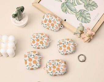 May include: White AirPod cases with a peach pattern are displayed. The cases have a rounded shape and a small loop for attaching a keychain. A framed botanical print and small figurines are also in the image.