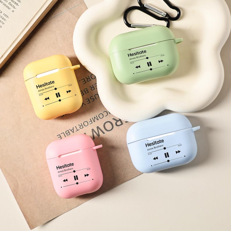 May include: Four colorful AirPod cases in yellow, green, pink, and blue. Each case has the text "Hesitate Jonas Brothers" with a music player graphic. A black carabiner clip is attached to one case.