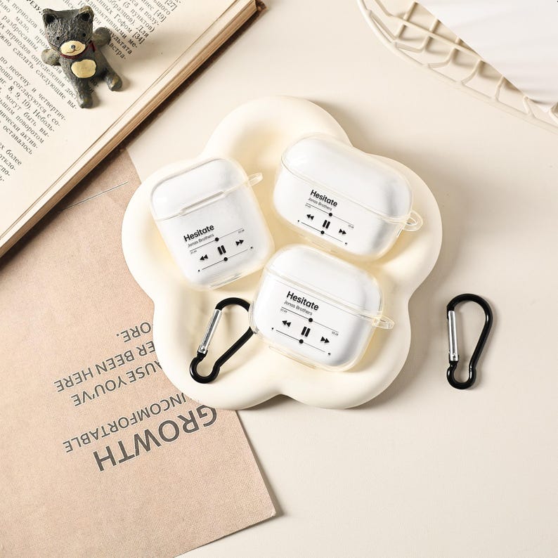 May include: Three white AirPod cases with the word "Hesitate" and a music player graphic printed on them. The cases are on a flower-shaped dish, with two black carabiner clips. A small bear figurine and a book are in the background.