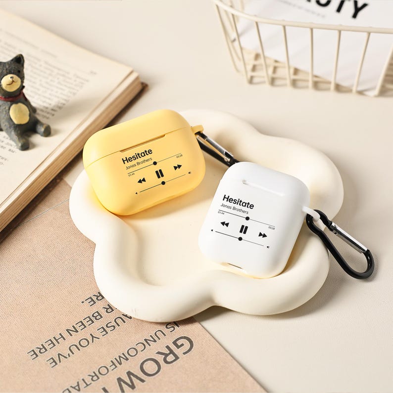 May include: Two wireless earbud cases, one yellow and one white, each with a black carabiner clip. The cases feature the text "Hesitate Jonas Brothers" and a music player graphic. They rest on a cream-coloured, cloud-shaped tray.