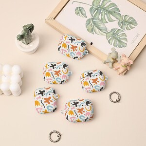 May include: Five white protective cases for wireless earbuds, each featuring a colorful abstract pattern of orange, pink, blue, and black shapes. The cases include a small side loop for attachment and are accompanied by two silver metal key rings.