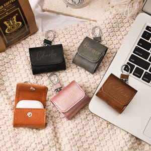 Puede incluir: Varios estuches de cuero para AirPod en negro, gris, marrón y rosa, cada uno con un llavero plateado. Los estuches están grabados con nombres. Un estuche está abierto, mostrando auriculares blancos.