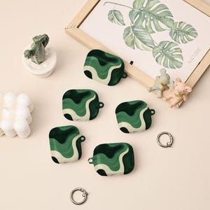 May include: Five green and white wave-patterned AirPod cases are shown. The cases feature a dark green base with lighter green and white wavy lines. A framed botanical print and small figurines are in the background. Two silver key rings are also visible.