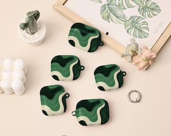 May include: Five green and white wave-patterned AirPod cases are displayed. The cases have a dark green base with lighter green and white wavy lines. A framed botanical print and small figurines are in the background. Two silver key rings are also visible.