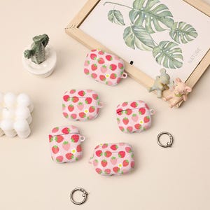 Könnte beinhalten: Rosa AirPod-Hüllen mit einem sich wiederholenden Erdbeermuster. Die Hüllen haben einen weißen Hintergrund mit roten Erdbeeren und grünen Blättern. Das Bild enthält auch einen gerahmten botanischen Druck, kleine Figuren und Schlüsselanhänger.