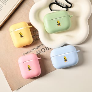Może przedstawiać: Cztery kolorowe etui na AirPods w kolorze żółtym, zielonym, różowym i niebieskim, każde z postacią z kreskówki. Etui ułożone są na białej, falistej tacy z czarnym karabińczykiem.
