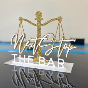 May include: A gold glitter scale of justice cake topper with the words "Next Stop THE BAR" in white script. The topper is on a white base, reflecting on a black surface.