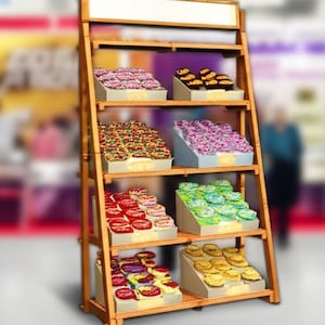Puede incluir: Un expositor de madera lleno de cupcakes con glaseados y decoraciones coloridas. Los cupcakes están dispuestos en estantes inclinados. El expositor tiene una estructura de madera clara y un letrero en blanco en la parte superior.