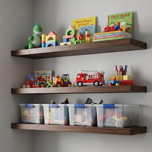 Puede incluir: Tres estantes de madera montados en una pared blanca, llenos de juguetes infantiles. Se ven un dinosaurio verde, bloques de colores, libros, vehículos de juguete y contenedores de plástico transparente con tapas azules. Los estantes son de madera oscura.