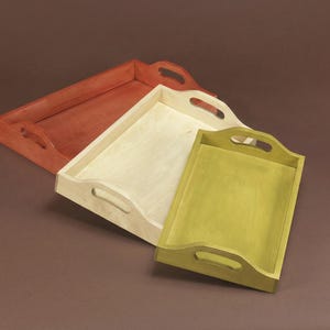 Puede incluir: Tres bandejas de madera para servir en tonos rojo, natural y verde. Cada bandeja tiene forma rectangular con asas curvas. Las bandejas están vacías y parecen hechas a mano.