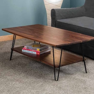 Puede incluir: Una mesa de centro de madera oscura con un estante inferior. La mesa tiene patas de horquilla negras y está sobre una alfombra gris claro. Hay algunos libros en el estante superior.