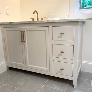 Puede incluir: Mueble de baño blanco con encimera de mármol y tres cajones. El mueble tiene dos puertas de armario con herrajes dorados. Los cajones también tienen herrajes dorados.